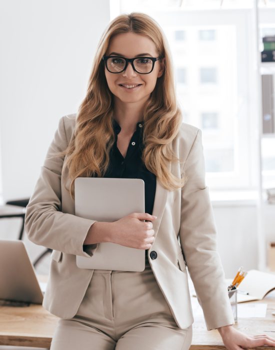 Confident business expert. Attractive young smiling woman in smart casual wear holding digital tablet and looking at camera while leaning on desk in creative office